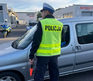 Zdjęcie w kolorze przedstawia na jezdni policjant kontroluje trzeźwość kierującego pojazdem osobowym.