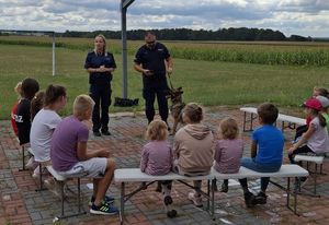 Zdjęcie w kolorze przedstawia dzieci siedzące na ławce na zewnątrz, za nimi para policjantów, którzy stoją obok siebie, policjant trzyma na smyczy psa. W tle łąka i niebo.