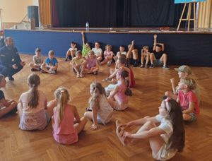 Zdjęcie w kolorze przedstawia dzieci siedzące dokoła na parkiecie wewnątrz sali miejskiego domu kultury w Oleśnie, obok nich kuca policjant.