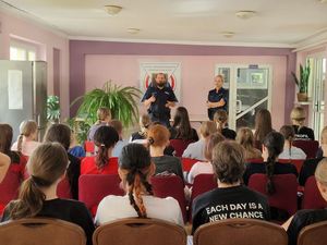 Zdjęcie w kolorze przedstawia dziewczęta siedzące w pomieszczeniu na krzesłach, na środku stoją policjant i obok po prawej stronie policjantka.