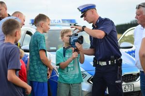 Zdjęcie w kolorze, przedstawia policjanta, który pokazuje obsługę miernika prędkości, przy funkcjonariuszu stoją młodzi chłopcy w wieku około 10 lat. Za nimi zaparkowany radiowóz.