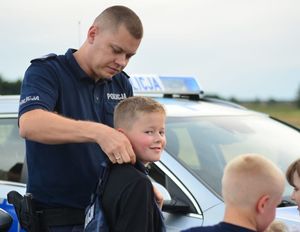 Zdjęcie w kolorze przedstawia policjanta, który zakłada chłopcu w wieku około 8 lat kamizelkę kuloodporną z napisem policja.