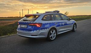 Zdjęcie w kolorze przedstawia policyjny pojazd na jezdni w tle za nim niebo rozświetlone zachodzącym słońcem.