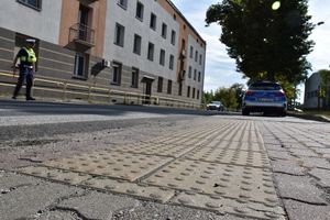 Zdjęcie w kolorze przedstawia rejon przejścia dla pieszych, po lewej policjant stoi na jezdni daje sygnał do zatrzymania nadjeżdżającemu pojazdowi koloru białego. po prawej zaparkowany radiowóz.