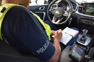 Zdjęcie w kolorze przedstawia policjanta który siedzi w radiowozie i  wypisuje blankiet mandatu karnego.
