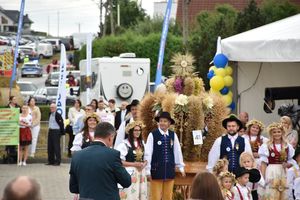 Zdjęcie w kolorze przedstawia uczetsników dożynek oraz osoby niosace koronę dożynkową  w strojach tradycyjnych ludowych. W tle  biały  namiot  w oddali radiowóz zabezpieczający imprezę.