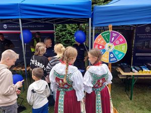 Zdjęcie w kolorze przedstawia dwie dziewczyny w strojach ludowych stojące przed policyjnym stoiskiem.