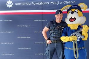 Zdjęcie w kolorze przedstawia policjanta pozującego do zdjęcia z maskotką Opolskiej Policji - Policusiem , który trzyma w łapach karabin AKMS. Oboje stoją na tle ściany z płótna z napisem Komenda Powiatowa Policji w Oleśnie.