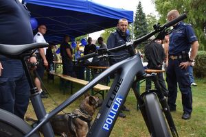 Zdjęcie w kolorze przedstawia fragment stoiska policyjnego z rowerami słuzbowymi oraz stojącym obok psem policyjnym, w tle uczestnicy imprezy odwiedzający stoisko.