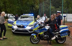 Zdjęcie w kolorze przedstawia młoda kobietę siedzącą na policyjnym motocyklu, tuz obok stoi policjant a dalej za nimi samochód.