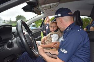 Zdjęcie w kolorze przedstawia policjanta wewnątrz radiowozu, który pokazuje dziecku konsole do włączania sygnałów.