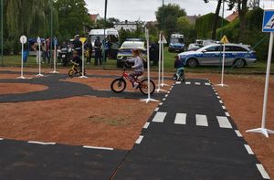 Zdjęcie w kolorze przedstawia miasteczko ruchu drogowego po którym jeżdżą najmłodsze dzieci na rowerkach.