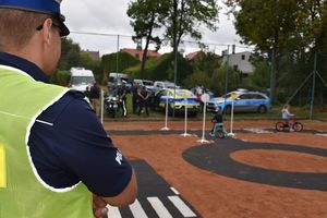 Zdjęcie w kolorze przedstawia miasteczko ruchu drogowego po którym jeżdżą najmłodsze dzieci na rowerkach na pierwszym planie stoi policjant w kamizelce odblaskowej.