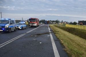 Zdjęcie w kolorze przedstawia jezdnię, na której po lewej stronie  stoją pojazdy policji i straży pożarnej z włączonymi światłami błyskowymi. Na prawym pasie fragmenty rozbitego samochodu osobowego.