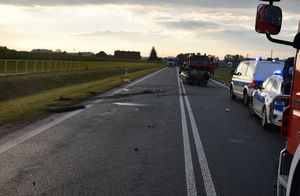 Zdjęcie w kolorze przedstawia jezdnię, na której po prawej stronie  stoją pojazdy policji i straży pożarnej z włączonymi światłami błyskowymi, za nimi rozbity pojazd leżący na dachu. Na lewym pasie fragmenty rozbitego samochodu osobowego.