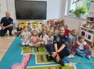 Zdjęcie w kolorze przedstawia przedszkolaków siedzących na podłodze wspólnie z policjantką.