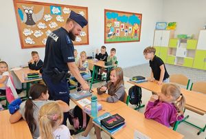 Zdjęcie w kolorze przedstawia policjanta stojącego pomiedzy ławkami w których siedzą uczniowie.