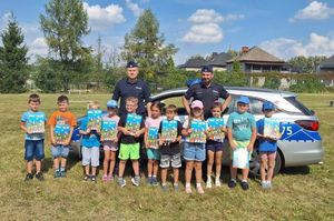 Zdjęcie w kolorze przedstawia uczniów na boisku szkolnym , trzymają w dłoniach książki pn.  Pierwszaki - pozują do zdjęcia z policjantem, który stoi pomiędzy nimi w tle błękitne niebo z białymi obłokami.