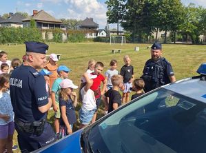 Zdjęcie w kolorze przedstawia uczniów na boisku szkolnym ,stoją przy radiowozie z policjantami.