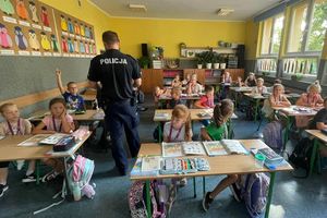 Zdjęcie w kolorze przedstawia policjanta stojącego pomiedzy ławkami w których siedzą uczniowie.