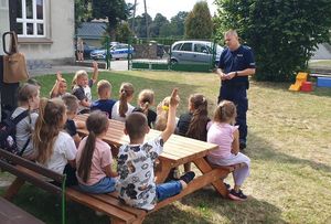 Zdjęcie w kolorze przedstawia policjanta stojącego na zewnątrz szkoły, uczniowie siedzą w ławce.