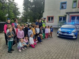 Zdjęcie w kolorze przedstawia grupę dzieci przedszkolnych na zewnętrznym placu, dzieci pozują do zdjęcia z policjantami obok radiowozu. W tle budynki i roślinność.