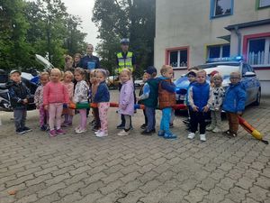 Zdjęcie w kolorze przedstawia grupę dzieci przedszkolnych na zewnętrznym placu, dzieci pozują do zdjęcia z policjantami obok radiowozu. W tle budynki i roślinność.