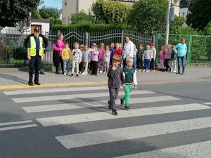 Zdjęcie w kolorze przedstawia grupę dzieci przedszkolnych stojących na chodniku, dwoje z nich przechodzi po pasach w towarzystwie policjantki.