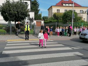 Zdjęcie w kolorze przedstawia grupę dzieci przedszkolnych stojących na chodniku, dwoje z nich przechodzi po pasach w towarzystwie policjantki.