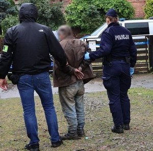 Zdjęcie w kolorze przedstawia parę policjantów prowadzących mężczyznę, który ma założone kajdanki na ręce trzymane z tyłu.W tle brama wjazdowa , za nią radiowóz.