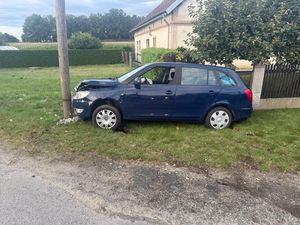 Zdjęcie w kolorze przedstawia granatowy samochód zaparkowany przy ogrodzeniu. Samochód posiada uszkodzony przedni zderzak i pokrywę silnika.