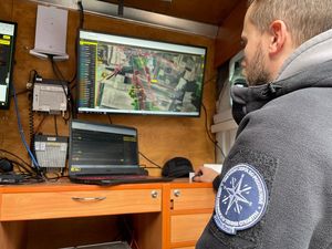 Zdjęcie w kolorze przedstawia wnętrze pojazdu z monitorami na których wyświetlane są mapy. Przed monitorem stoi mężczyzna w zieklonej kurtce z grupy poszukiwawczej.
