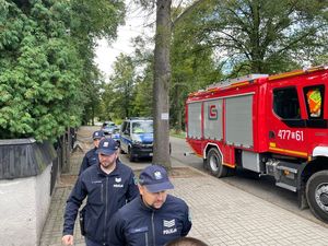 Zdjęcie w kolorze przedstawia policjantów idących chodnikiem obok zaparkowanych wozów straży pożarnej.