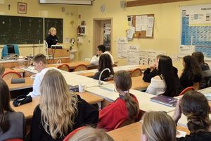 Zdjęcie w kolorze przedstawia uczniów siedzących w klasie  przed tablicą stoi policjantka.