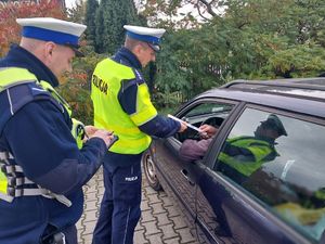 Zdjęcie w kolorze przedstawia policjantów podchodzących do kontrolowanego samochodu osobowego. Pierwszy z funkcjonariuszy wręcza kierującemu odblask.