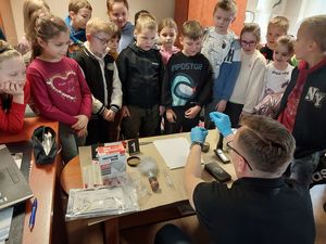 Zdjęcie w kolorze przedstawia grupę dzieci w pomieszczeniu technika kryminalistyki podczas pokazu ujawniania i utrwalania śladów.