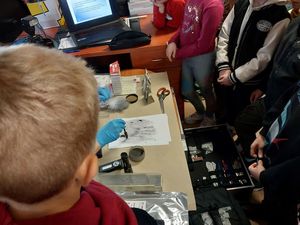 Zdjęcie w kolorze przedstawia grupę dzieci w pomieszczeniu technika kryminalistyki podczas pokazu ujawniania i utrwalania śladów.