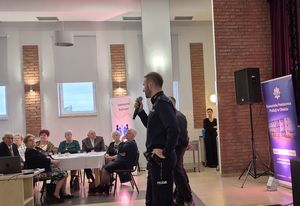 Zdjęcie w kolorze przedstawia policjanta trzymającego w dłoni mikrofon, za nim siedzący za stołem seniorzy.