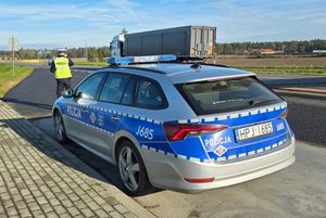Zdjęcie w kolorze przedstawia radiowóz stojący na miejscu kontroli drogowej, za nim stoi  policjant w tle przejeżdżający pojazd ciężarowy,