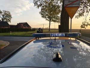 Zdjęcie w kolorze, przedstawia dach radiowozu, za nim stojący na drodze policjant z miernikiem prędkości w dłoniach.