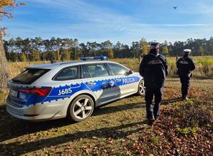 Zdjęcie w kolorze przedstawia dwóch policjantów obok radiowozu, w oddali na niebie lecący dron.