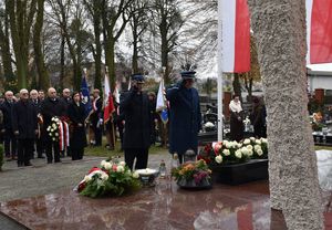 Zdjęcie w kolorze przedstawia delegację służb mundurowych - policji i straży pozarnej podczas składania wieńca z biało czerwonych kwiatów na pomniku lotników polskich.