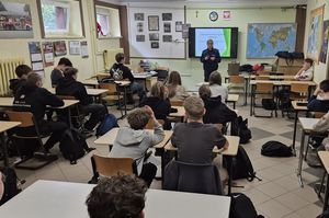 Zdjęcie w kolorze przedstawia klasę z uczniami siedzącymi w ławkach, na środku przy tablicy stoi policjantka.