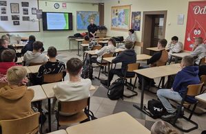 Zdjęcie w kolorze przedstawia klasę z uczniami siedzącymi w ławkach, na środku przy tablicy stoi policjantka.