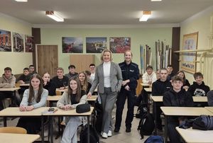 Zdjęcie w kolorze przedstawia klasę z uczniami siedzącymi w ławkach, na środku pomiędzy ławkami stoją dwie kobiety : policjantka i pani rzecznik konsumentów.