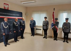 Zdjęcie w kolorze przedstawia panów komendantów na tle ściany z płótna z napisem Komenda Powiatowa Policji w Oleśnie, po prawej Poczet Sztandarowy.