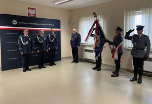 Zdjęcie w kolorze przedstawia panów komendantów na tle ściany z płótna z napisem Komenda Powiatowa Policji w Oleśnie, po prawej Poczet Sztandarowy.