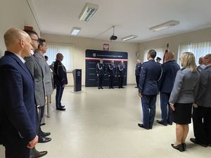 Zdjęcie w kolorze przedstawia zgromadzonych gości na pierwszym planie po lewej oraz policjantów po prawej, na dalszym planie panów komendantów na tle ściany z płótna z napisem Komenda Powiatowa Policji w Oleśnie, po prawej Poczet Sztandarowy.