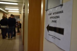 Zdjęcie w kolorze przedstawia, Napis na kartce przyklejonej do ściany ze strzałką - punkt wydawania zaopatrzenia.