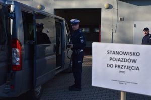 Zdjęcie w kolorze przedstawia stanowisko do przyjęcia pojazdów obsługiwane przez policjanta, który fotografuje pojazd typu bus.
